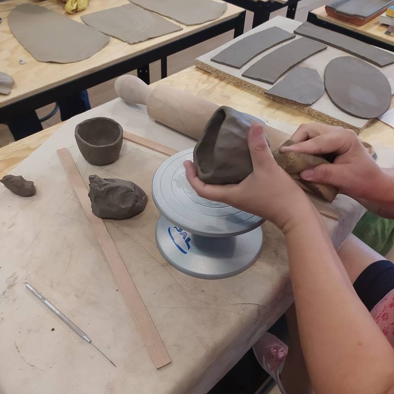 Aluna a trabalhar à roda de oleiro, com as estantes do atelier ao fundo
